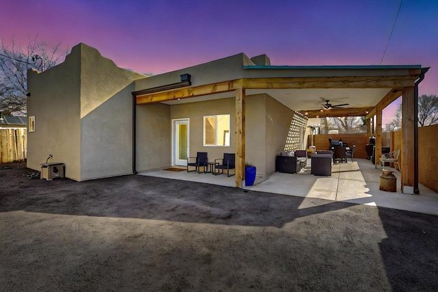 back of house at dusk with ceiling fan, stucco siding, fence, and a patio