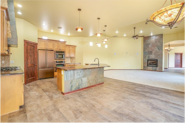 kitchen featuring decorative backsplash, ceiling fan with notable chandelier, a tile fireplace, appliances with stainless steel finishes, and a kitchen island with sink