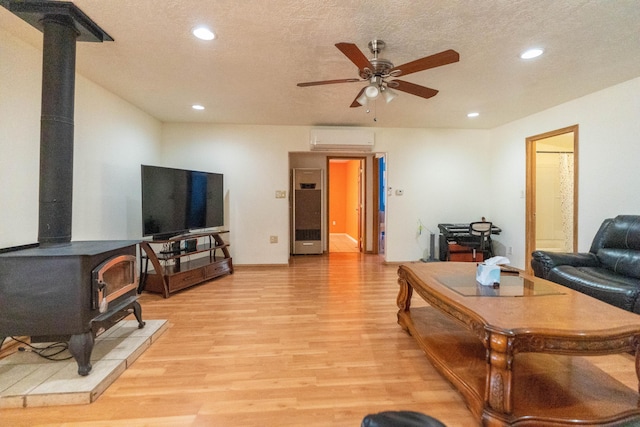 living room featuring ceiling fan, a textured ceiling, a wall unit AC, light wood-type flooring, and a wood stove