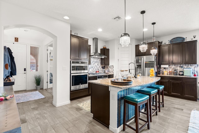 kitchen with arched walkways, visible vents, stainless steel appliances, dark brown cabinets, and wall chimney range hood