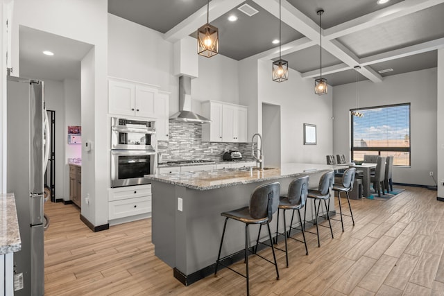 kitchen featuring coffered ceiling, light wood-style floors, appliances with stainless steel finishes, wall chimney range hood, and a sink