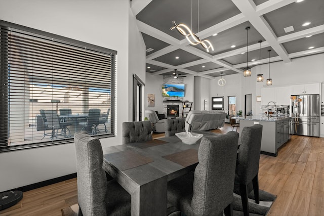 dining space with light wood finished floors, coffered ceiling, a towering ceiling, a stone fireplace, and beam ceiling