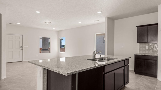 kitchen with a textured ceiling, a sink, light stone countertops, tasteful backsplash, and a center island with sink