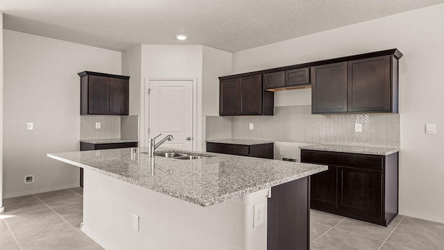 kitchen featuring decorative backsplash, an island with sink, light stone countertops, dark brown cabinets, and a sink