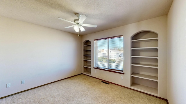 carpeted spare room featuring a textured ceiling, a ceiling fan, baseboards, visible vents, and built in features