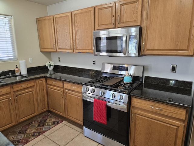 kitchen featuring light tile patterned floors, dark stone counters, brown cabinets, stainless steel appliances, and a sink