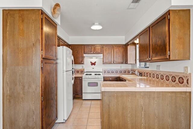 kitchen featuring visible vents, under cabinet range hood, white appliances, a peninsula, and light tile patterned floors