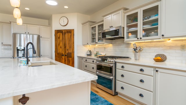 kitchen with light wood finished floors, decorative backsplash, glass insert cabinets, stainless steel appliances, and a sink