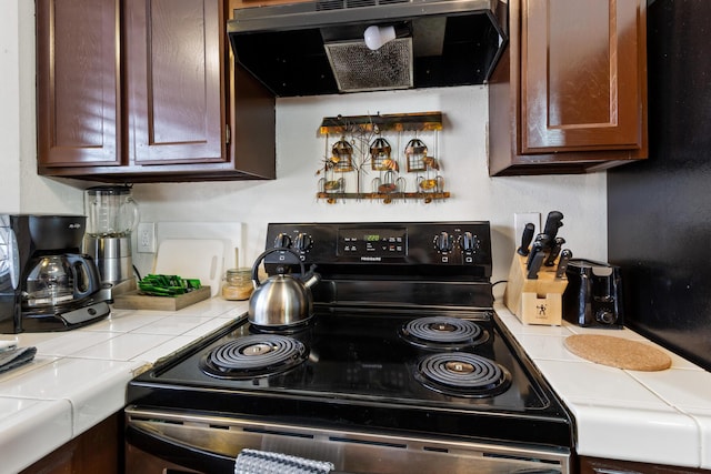 kitchen featuring black range with electric cooktop, tile countertops, dark brown cabinets, and exhaust hood