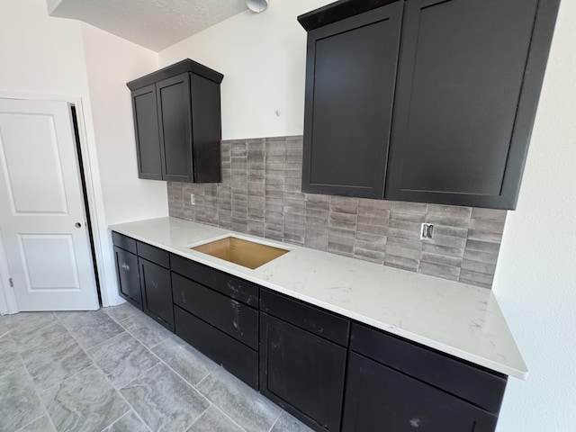 kitchen featuring a textured ceiling, dark cabinets, a sink, backsplash, and light stone countertops