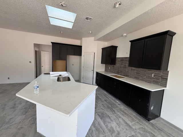 kitchen featuring a skylight, visible vents, dark cabinetry, decorative backsplash, and an island with sink