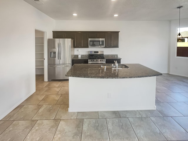 kitchen featuring dark brown cabinetry, an island with sink, appliances with stainless steel finishes, dark stone countertops, and a sink