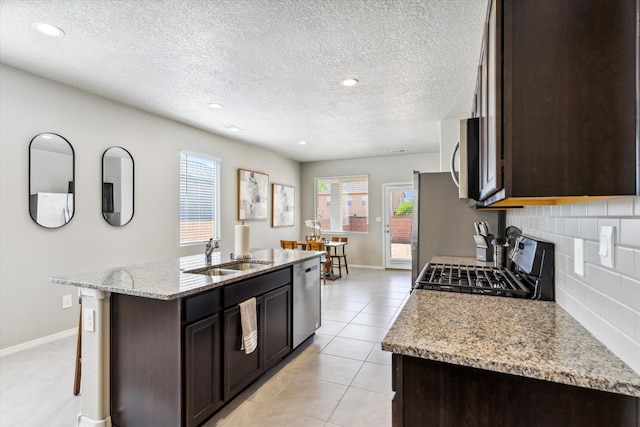 kitchen featuring a kitchen island with sink, dark brown cabinetry, stainless steel appliances, a sink, and tasteful backsplash