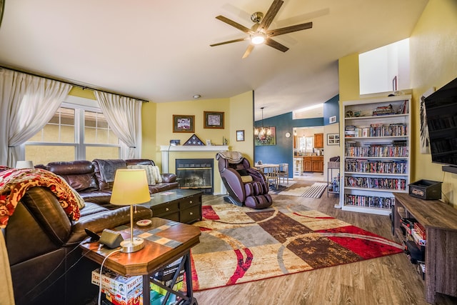 living room with ceiling fan with notable chandelier, lofted ceiling, a glass covered fireplace, and wood finished floors