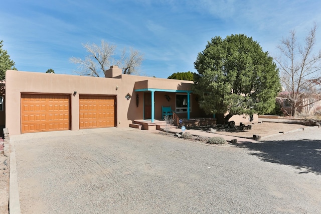 pueblo-style house featuring gravel driveway, a porch, a chimney, stucco siding, and a garage