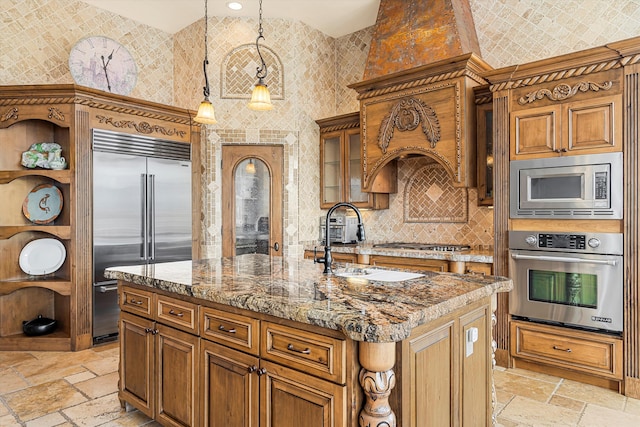 kitchen with stone tile flooring, brown cabinets, an island with sink, and built in appliances