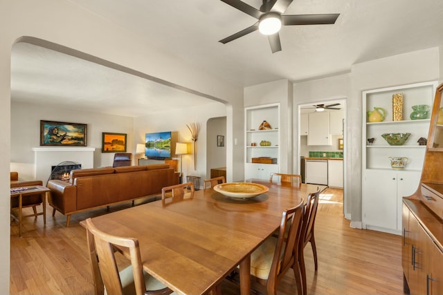 dining area featuring built in shelves, arched walkways, a warm lit fireplace, and light wood-style flooring