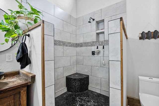 bathroom with a textured wall, toilet, vanity, and a tile shower