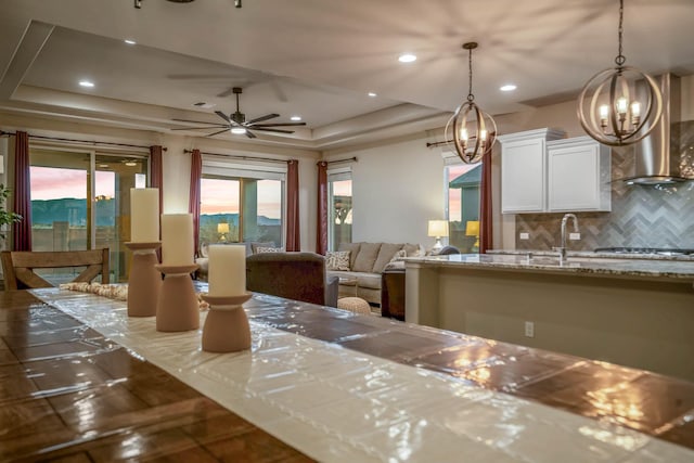 kitchen with a tray ceiling, decorative backsplash, white cabinets, wall chimney range hood, and open floor plan