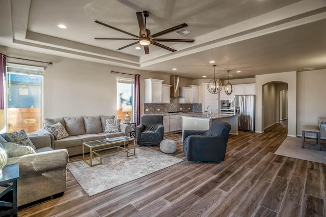 living area featuring ceiling fan with notable chandelier, a raised ceiling, dark wood-style floors, and arched walkways