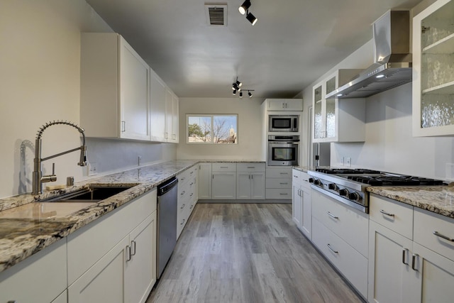 kitchen featuring a sink, light wood-style floors, appliances with stainless steel finishes, wall chimney exhaust hood, and glass insert cabinets