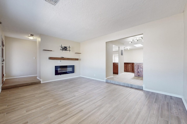 unfurnished living room with baseboards, visible vents, a glass covered fireplace, a textured ceiling, and light wood-type flooring