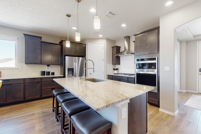 kitchen with light stone counters, appliances with stainless steel finishes, a sink, wall chimney range hood, and dark brown cabinets