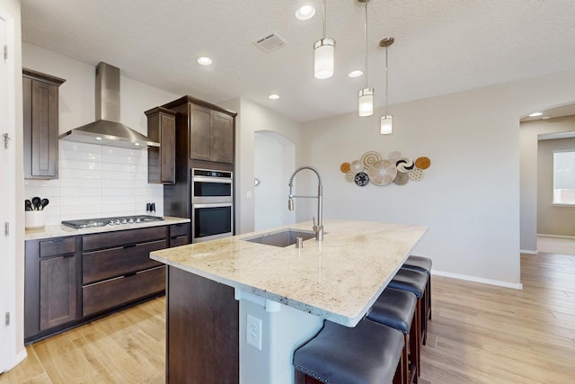 kitchen with arched walkways, a sink, appliances with stainless steel finishes, wall chimney exhaust hood, and an island with sink