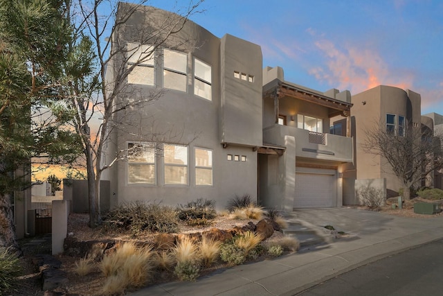 pueblo-style house with an attached garage, a balcony, driveway, and stucco siding