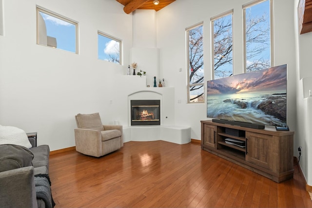 living room with a high ceiling, a glass covered fireplace, beamed ceiling, baseboards, and hardwood / wood-style flooring