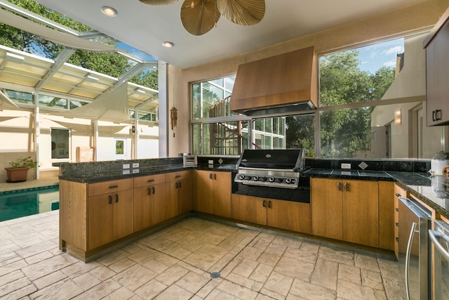 kitchen featuring stone tile floors, custom exhaust hood, brown cabinetry, ceiling fan, and a peninsula