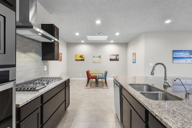 kitchen featuring light stone countertops, a sink, appliances with stainless steel finishes, wall chimney exhaust hood, and tasteful backsplash