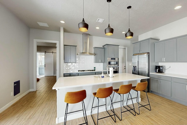 kitchen with stainless steel appliances, gray cabinetry, a barn door, wall chimney exhaust hood, and backsplash