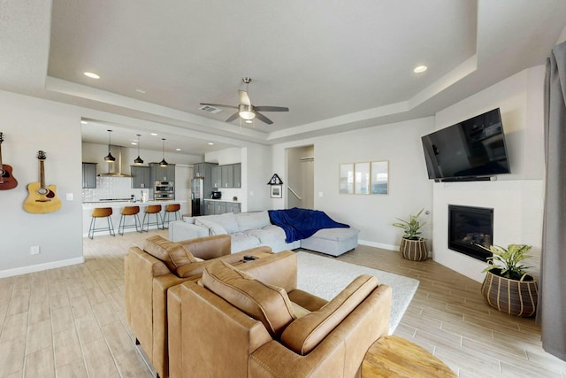 living room with a ceiling fan, baseboards, wood finish floors, a tray ceiling, and a glass covered fireplace