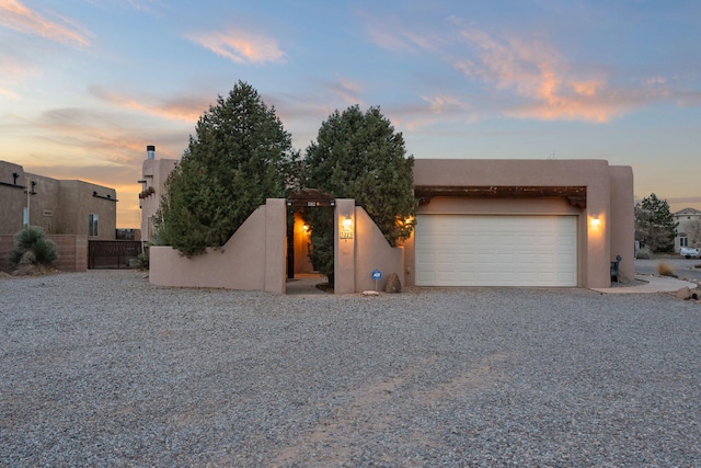 pueblo revival-style home featuring stucco siding, a gate, fence, gravel driveway, and an attached garage