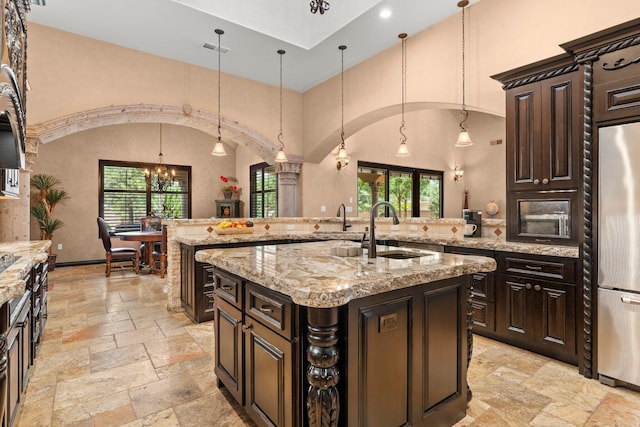 kitchen with an island with sink, stone tile floors, arched walkways, stainless steel fridge, and a sink