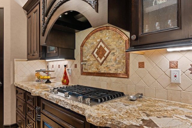 kitchen featuring light stone counters, stainless steel gas cooktop, dark brown cabinets, glass insert cabinets, and backsplash