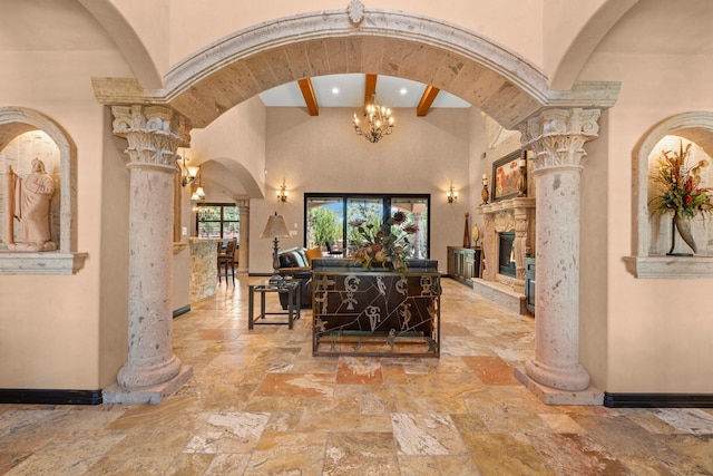 corridor featuring stone tile floors, a chandelier, decorative columns, baseboards, and a towering ceiling