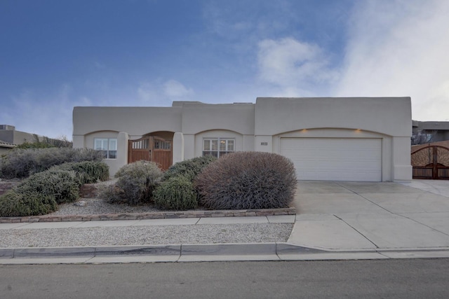 pueblo-style home with a gate, an attached garage, driveway, and stucco siding