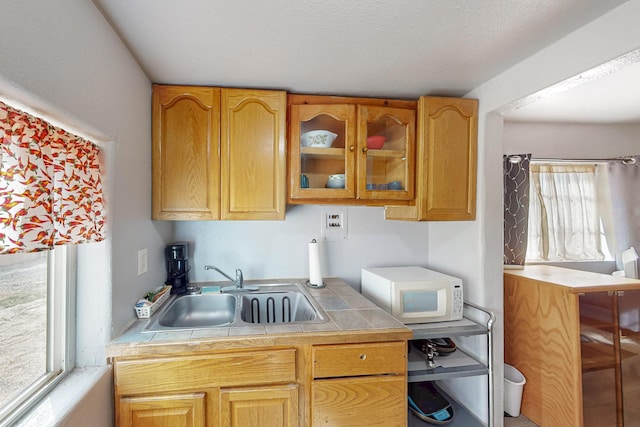 kitchen with tile countertops, white microwave, a sink, glass insert cabinets, and a textured ceiling