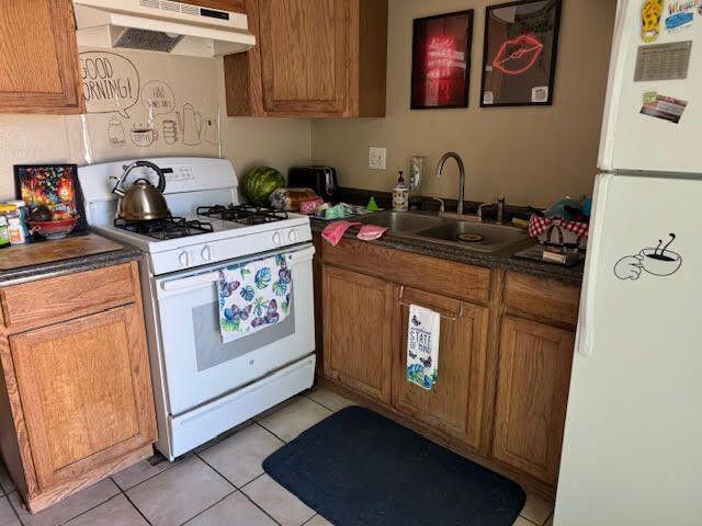 kitchen with white appliances, light tile patterned floors, brown cabinetry, a sink, and under cabinet range hood