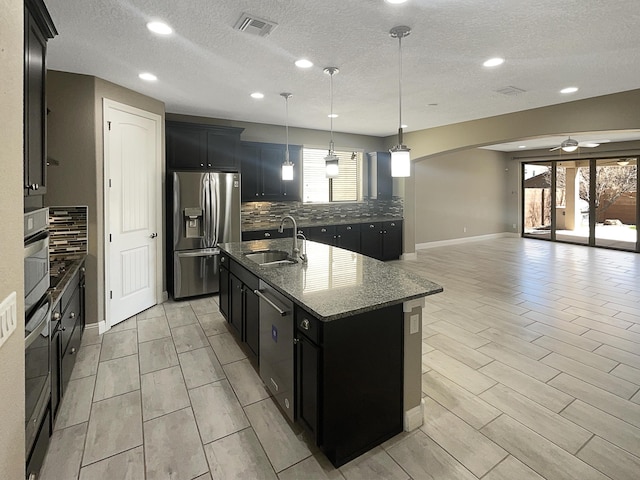 kitchen with a kitchen island with sink, a sink, dark cabinetry, appliances with stainless steel finishes, and decorative backsplash