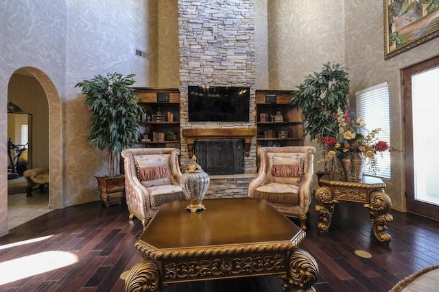 living room with arched walkways, a stone fireplace, wood finished floors, and a towering ceiling