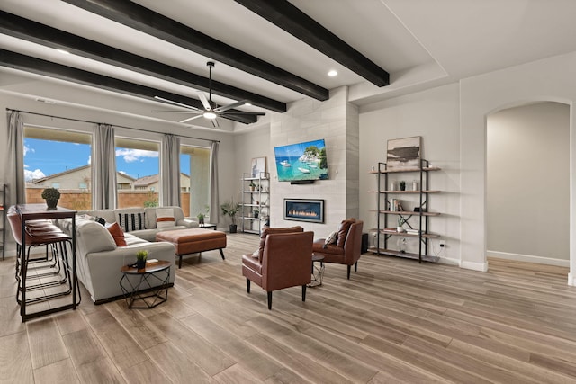 living room with arched walkways, beam ceiling, a tile fireplace, light wood-type flooring, and a ceiling fan
