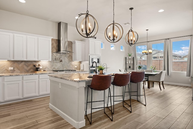 kitchen with a kitchen island with sink, an inviting chandelier, light wood-type flooring, backsplash, and wall chimney range hood