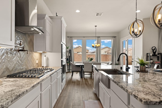 kitchen featuring wall chimney range hood, a chandelier, appliances with stainless steel finishes, white cabinetry, and a sink