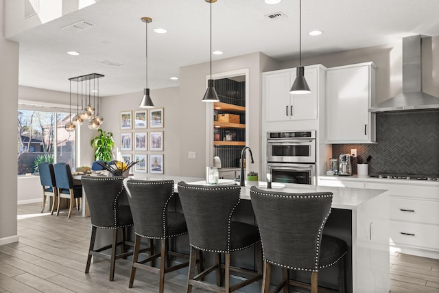 kitchen featuring backsplash, double oven, light wood-type flooring, black cooktop, and wall chimney exhaust hood