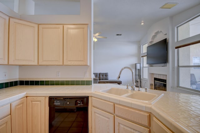 kitchen with a ceiling fan, light brown cabinetry, a sink, black dishwasher, and a fireplace