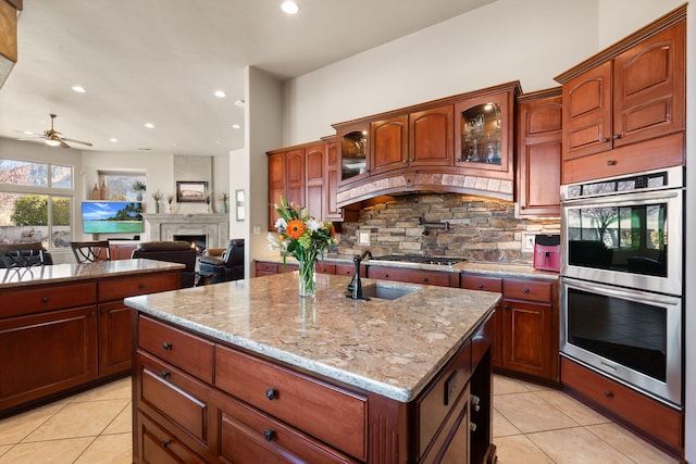 kitchen featuring stainless steel double oven, an island with sink, a sink, backsplash, and a large fireplace