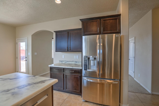 kitchen featuring stainless steel fridge, arched walkways, dark brown cabinetry, light tile patterned flooring, and light countertops